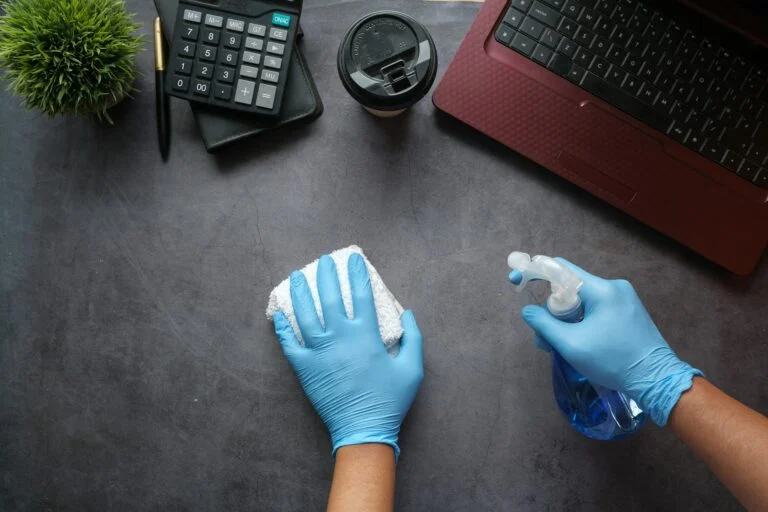 Gloved hands sanitising an office desk with spray and cloth